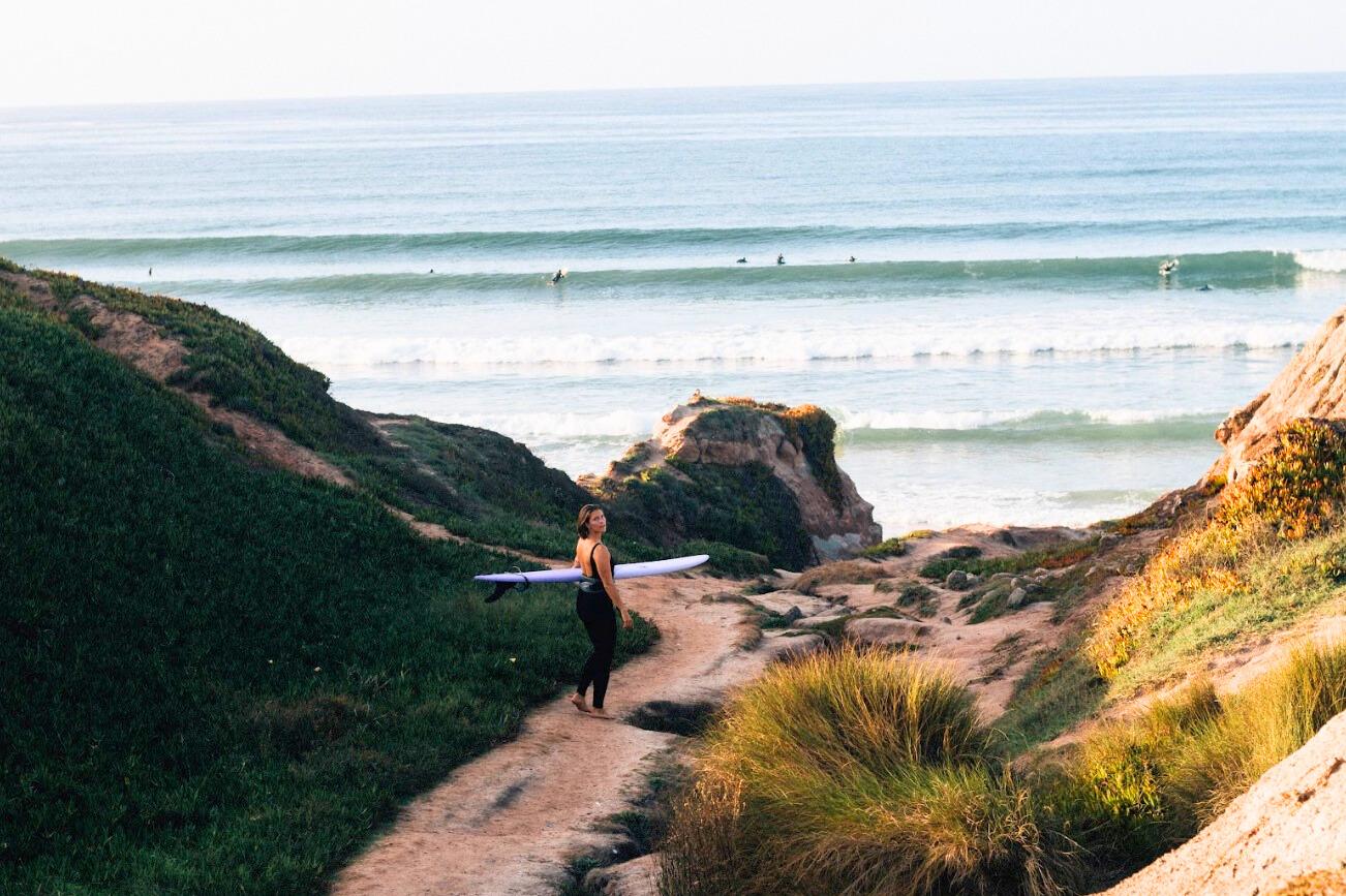 En surfare går längs en stig mot havet i Baleal, Portugal, med vågor i bakgrunden
